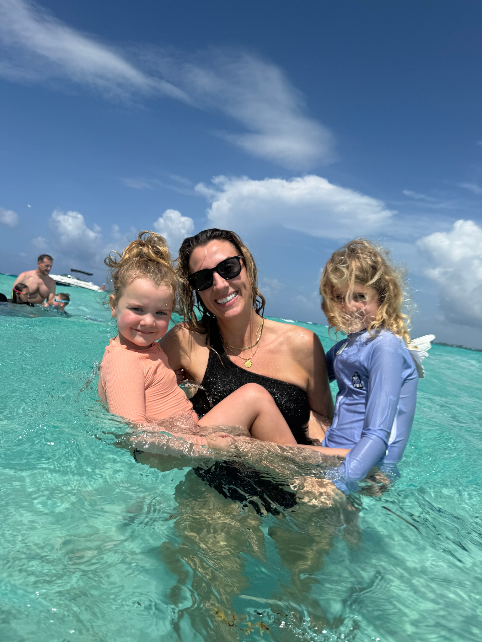 A parent holding two young children in calm, shallow turquoise water during a family outing in Grand Cayman, a boat in the background.