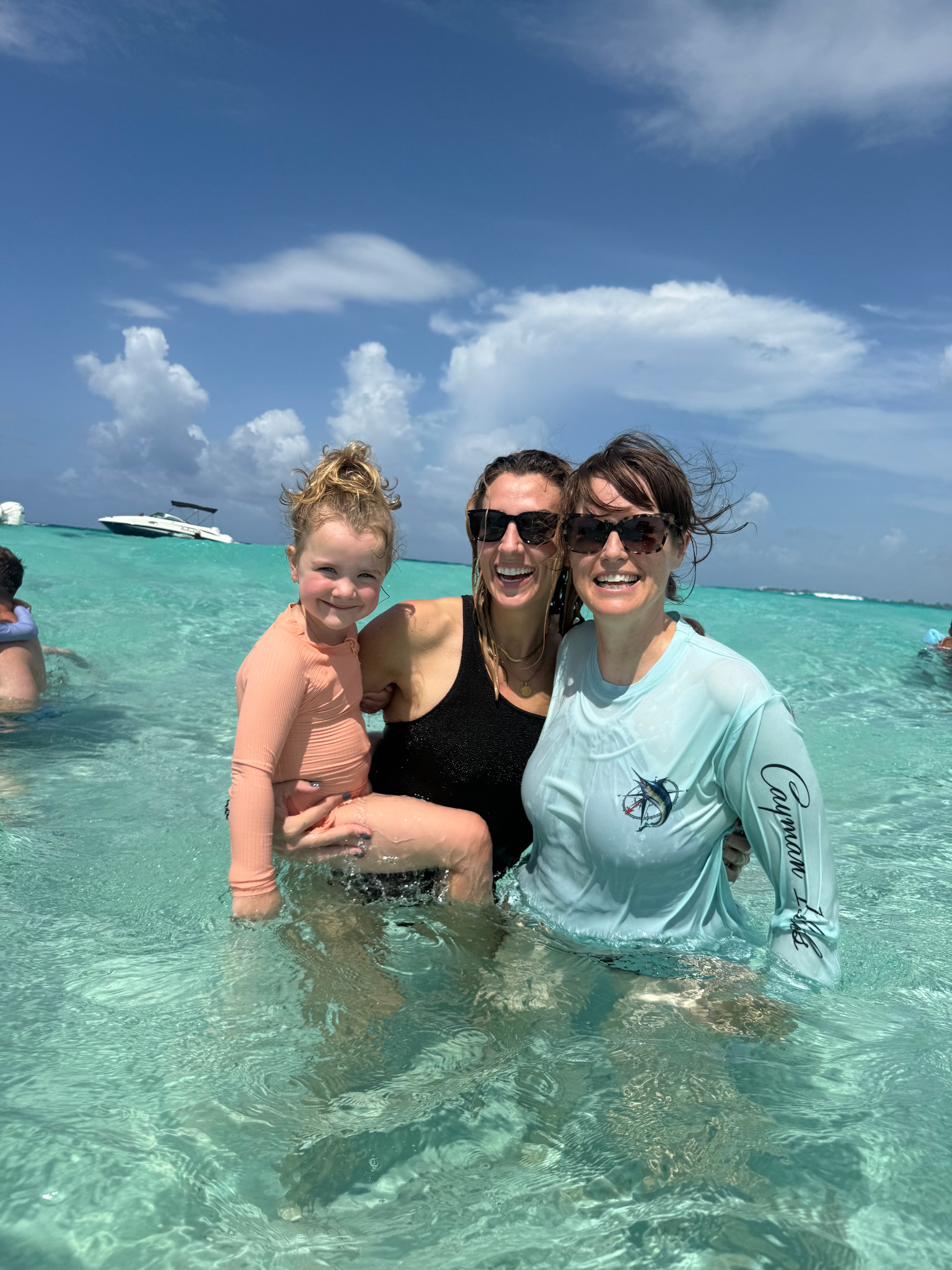 Two women and a young child smiling together in calm, shallow turquoise water during a family outing in Grand Cayman, a boat in the background.