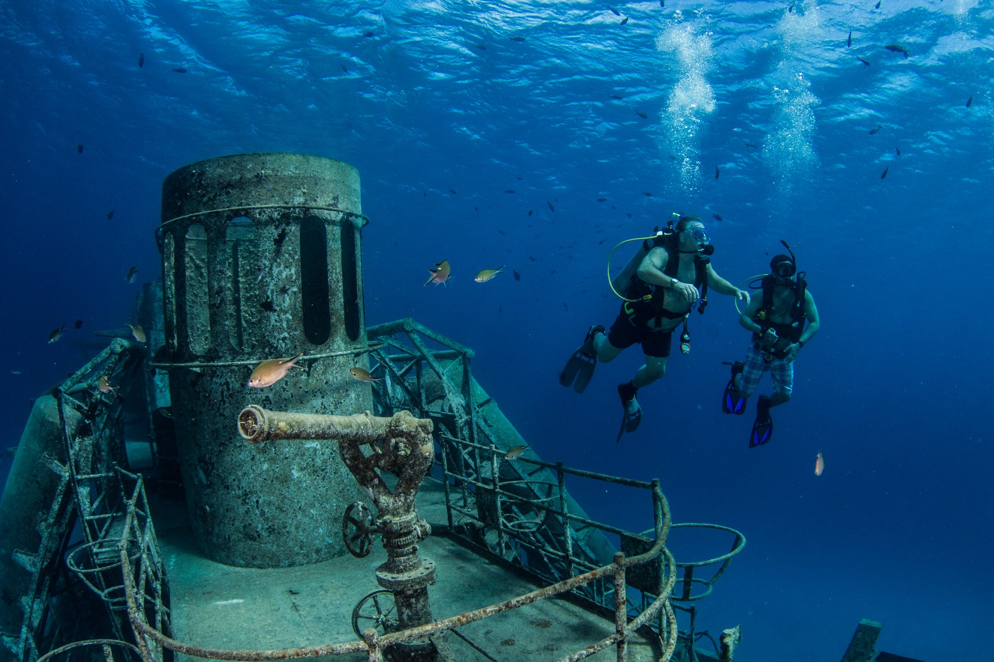 Two divers swimming above the open decks of the Kittiwake wreck in bright blue Grand Cayman water, with reef fish around the structure