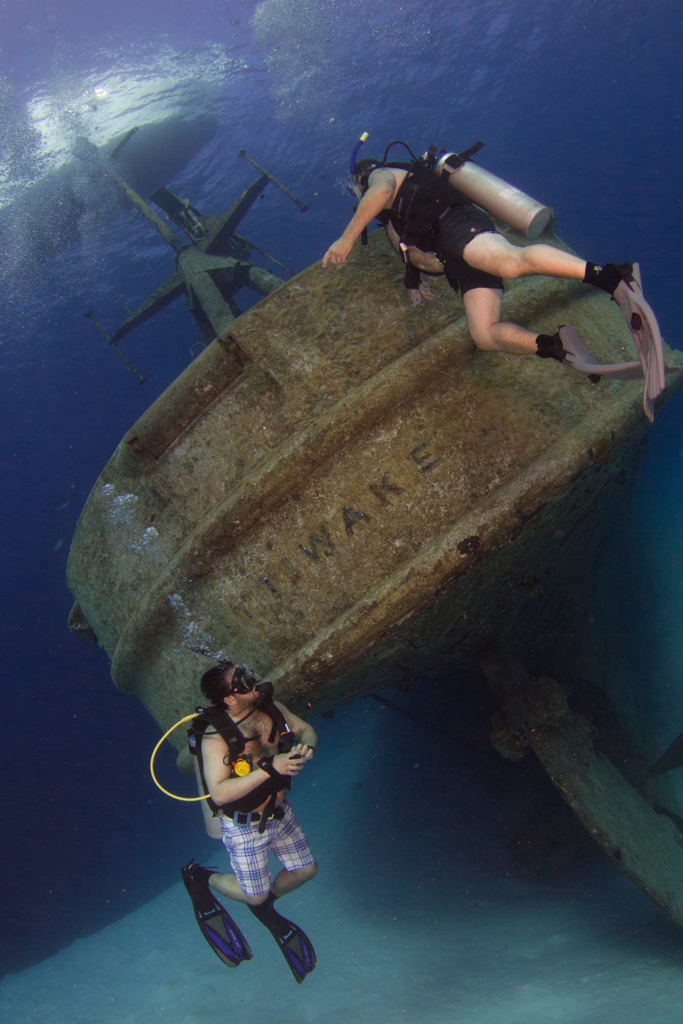 Two divers alongside the towering stern of the Kittiwake, showing the scale of the wreck against the white sand below