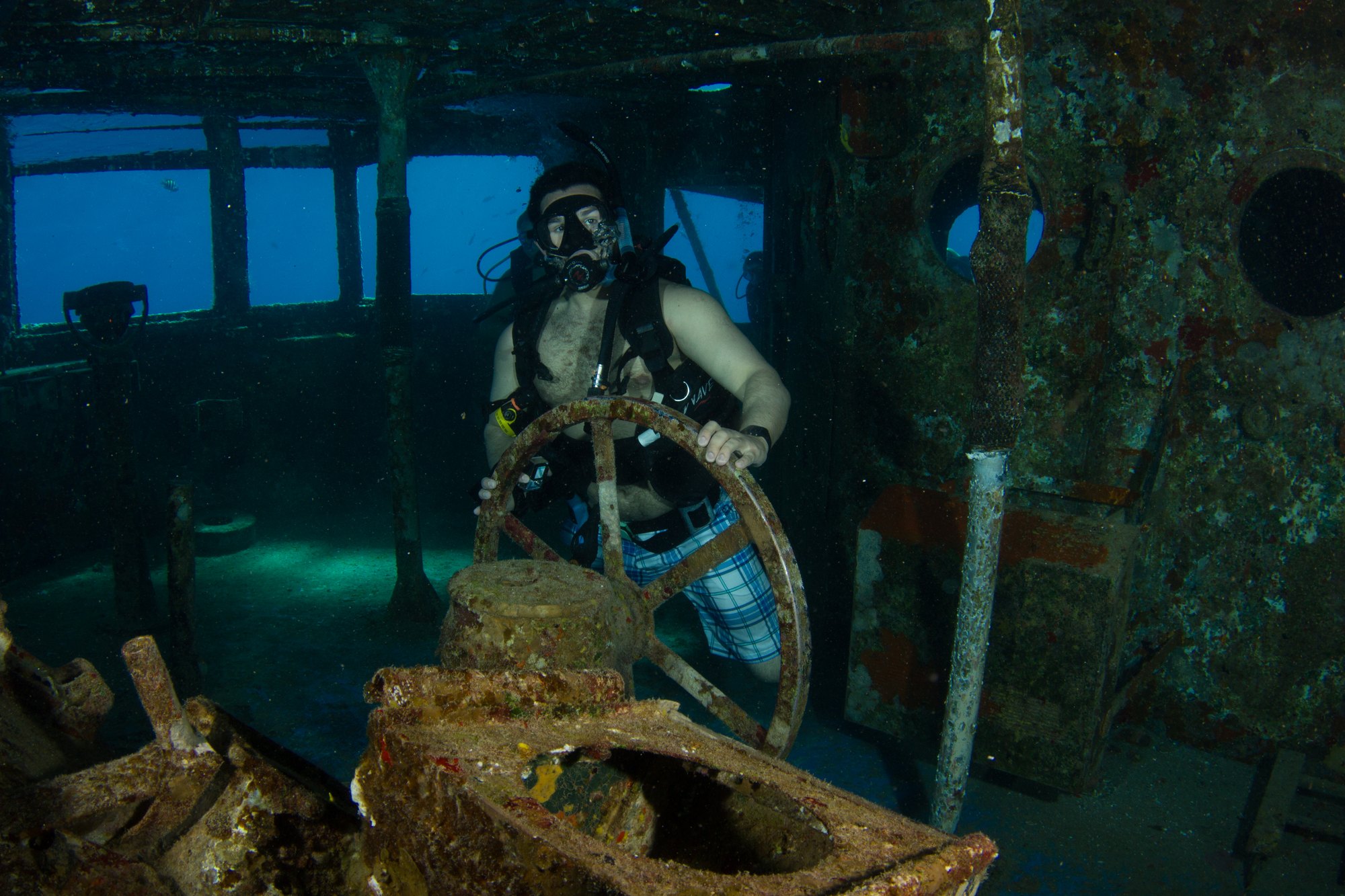 A diver at the ship's wheel inside the Kittiwake's wheelhouse, with daylight coming through the large open window openings on either side