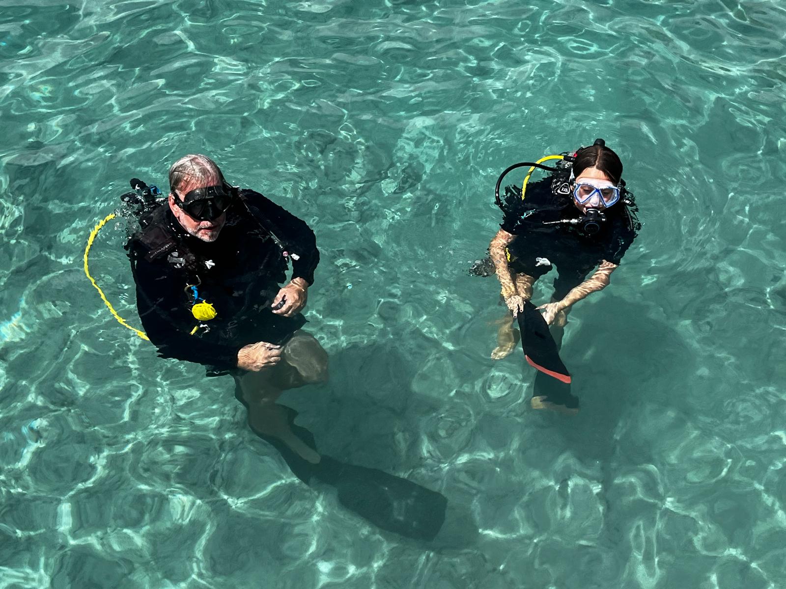 A DNS Diving instructor beside a young diver in gear, standing together in clear waist-deep turquoise water